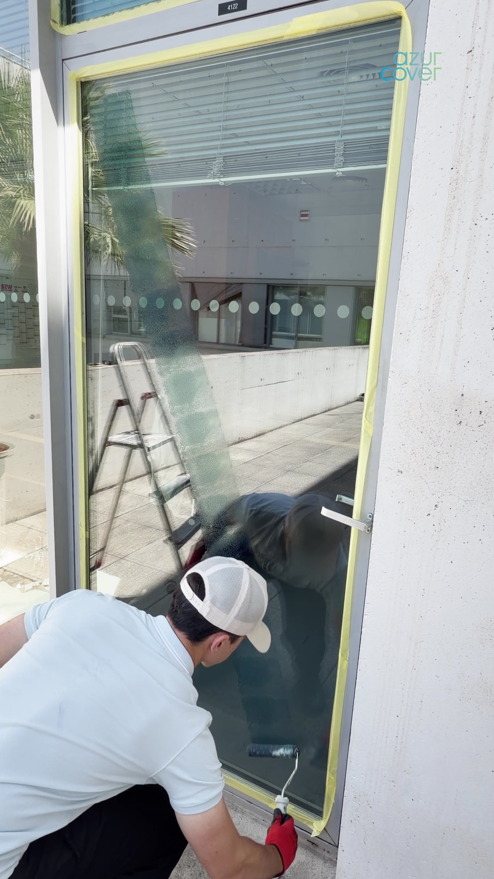 Technicien en gants rouges appliquant au rouleau un vernis transparent Azur Reflect sur une grande baie vitrée, châssis protégé par un ruban jaune.